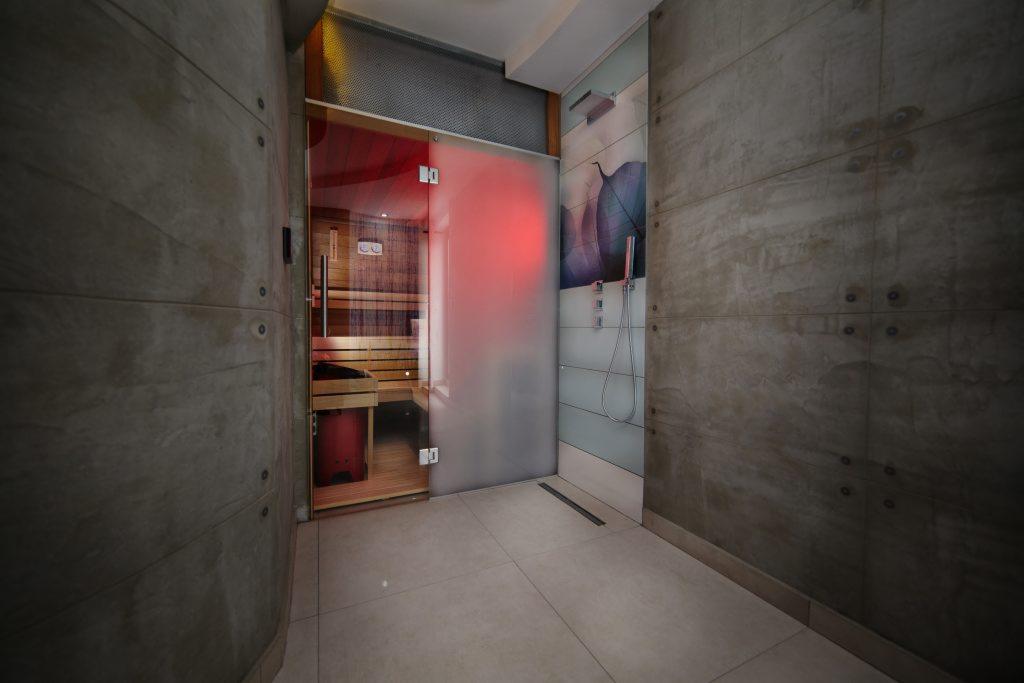 Nowoczesna łazienka z sauną i kabiną prysznicową, wykończona płytami betonowymi, z widocznym oświetleniem LED w saunie.