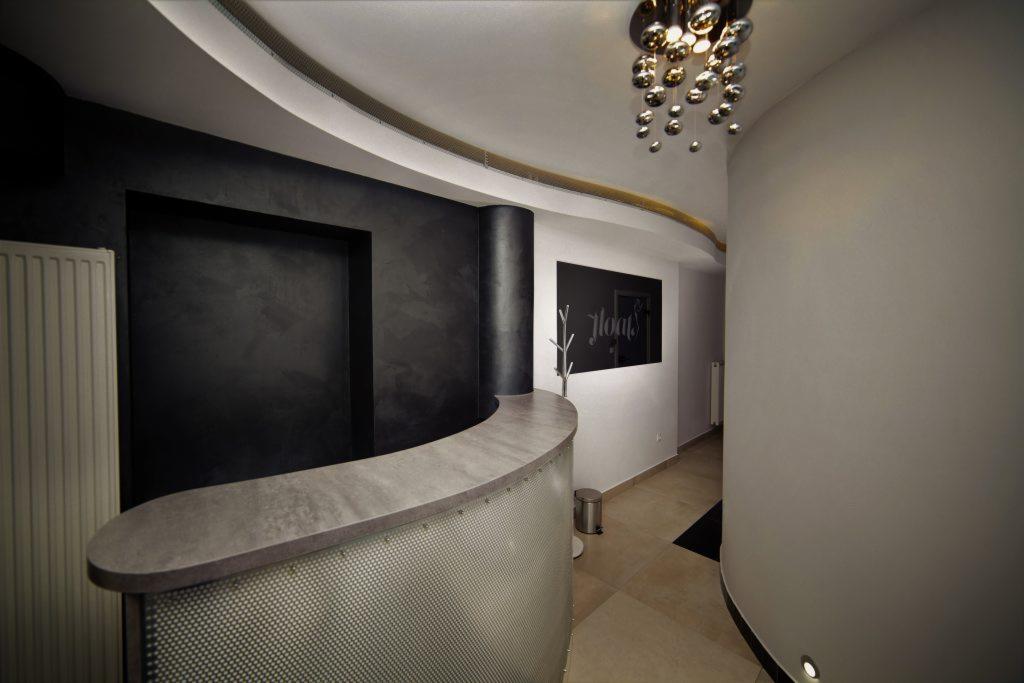 Nowoczesne wnętrze z zaokrągloną ladą recepcyjną wykończoną perforowaną blachą, ciemna ściana z dekoracyjną fakturą tynku, designerska lampa sufitowa i tablica z napisem.