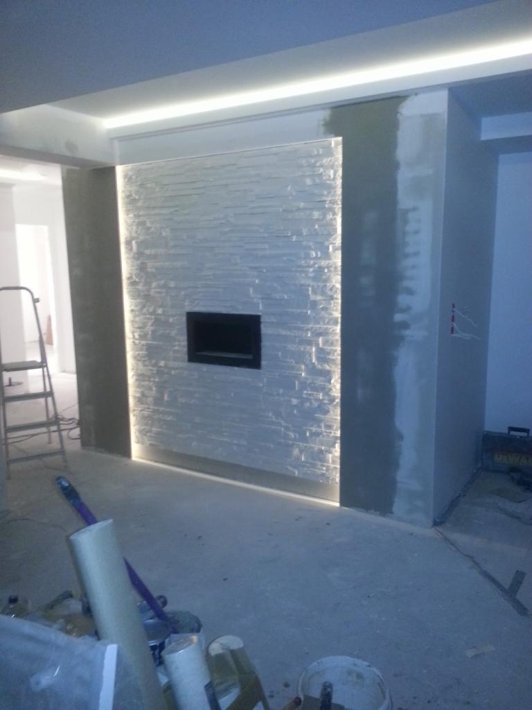 Ściana z dekoracyjnej białej cegły z wnęką na kominek, podświetlona listwami LED, w trakcie prac wykończeniowych. Widoczne ślady szpachlowania na ścianach bocznych i narzędzia na podłodze.