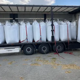 Ciężarówka marki Wielton załadowana workami z materiałem opałowym zabezpieczonymi czerwonymi pasami transportowymi, na ziemi rozsypane resztki materiału.