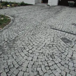 Podjazd wyłożony szarą kostką granitową w okrągły wzór, z widocznym odwodnieniem liniowym i fragmentem budynku w tle.
