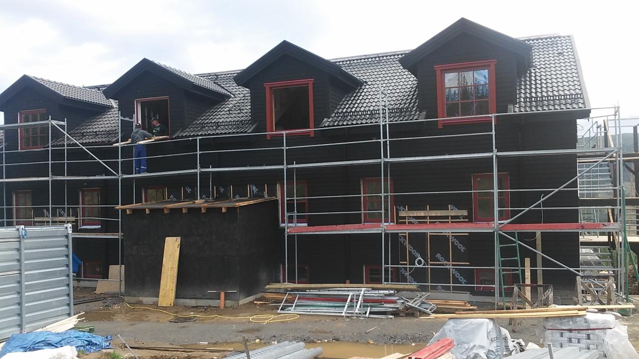 Montaż stolarki okiennej w budynku z ciemną elewacją, widoczne rusztowania i dwóch pracowników.