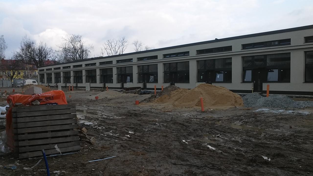 Budynek w trakcie budowy, widoczne nowe okna z czarnymi ramami, sterty piasku i gruzu na błotnistym placu budowy, paleta z materiałami budowlanymi w pierwszym planie.