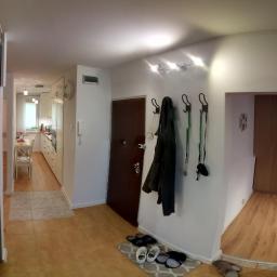 ExigoBud - Jasne wnętrze mieszkania z panelami podłogowymi, widok na przedpokój z wieszakami, brązowymi drzwiami i fragment kuchni w tle.