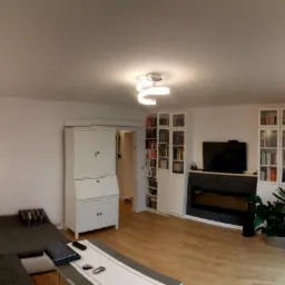 Jasny salon z drewnianą podłogą, białymi ścianami, narożną sofą, biurkiem sekretarzykiem i regałami na książki po bokach telewizora