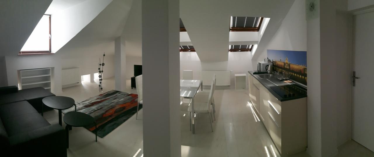 Jasne, przestronne poddasze z aneksem kuchennym, białymi ścianami i podłogą, oświetlone przez okna dachowe, z widocznym salonem z czarną sofą i stolikami kawowymi oraz elementami dekoracyjnymi.
