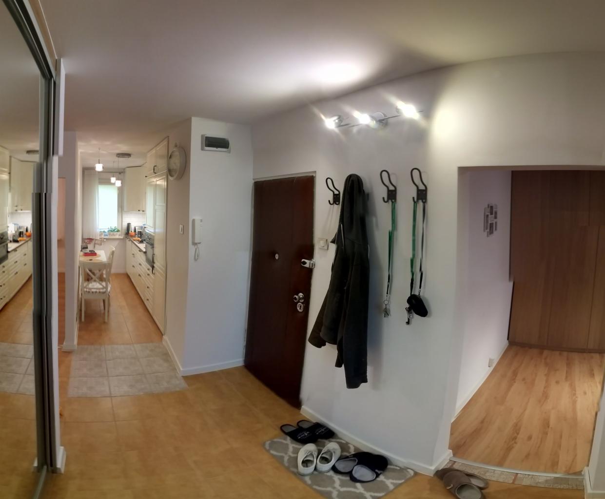 Jasne wnętrze mieszkania z panelami podłogowymi, widok na przedpokój z wieszakami, brązowymi drzwiami i fragment kuchni w tle.