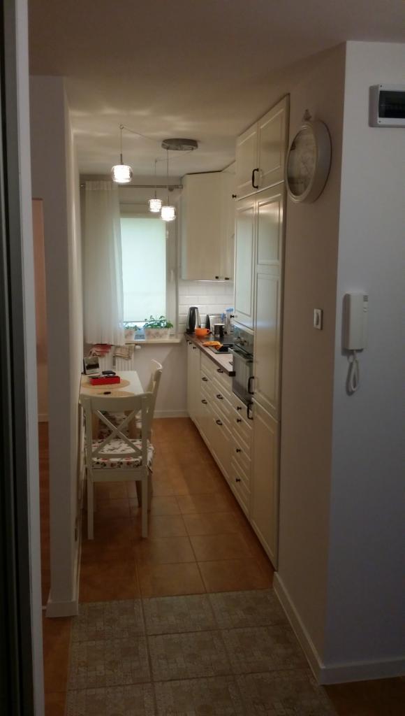 Wnętrze wąskiej kuchni z białymi meblami, ceramiczną podłogą i widoczną przestrzenią jadalną z krzesłem w stylu prowansalskim. Na ścianie wisi zegar i domofon.