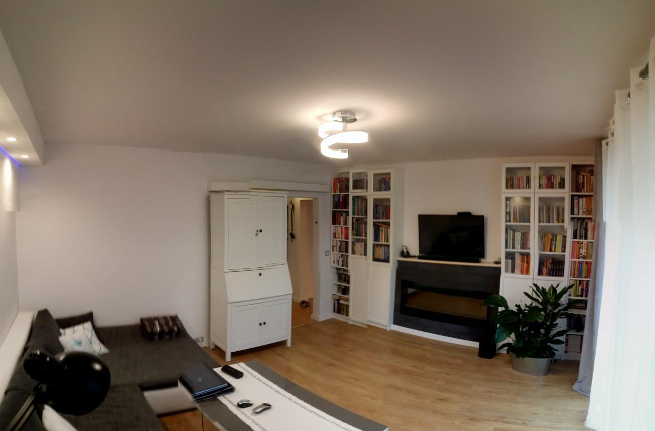 Jasny salon z drewnianą podłogą, białymi ścianami, narożną sofą, biurkiem sekretarzykiem i regałami na książki po bokach telewizora