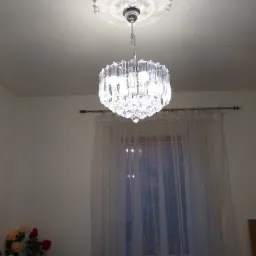 Kryształowy żyrandol zawieszony na łańcuchu pod ozdobnym, okrągłym rozetą sufitową, w tle okno z firanką i fragment ściany z kwiatami w wazonie.