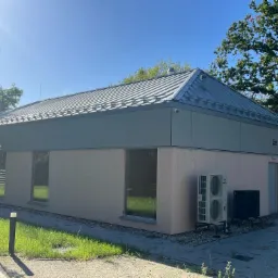 Nowoczesny budynek parterowy z jasną elewacją, szarym dachem i klimatyzacją, otoczony zielenią w słoneczny dzień. Widok z zewnątrz, prezentujący architekturę budynku.