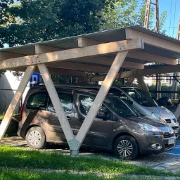Drewniana wiata parkingowa z blachy falistej, wsparta na konstrukcji z belek. Dwa zaparkowane samochody, jeden brązowy, drugi srebrny. Znak niebieski dla niepełnosprawnych.