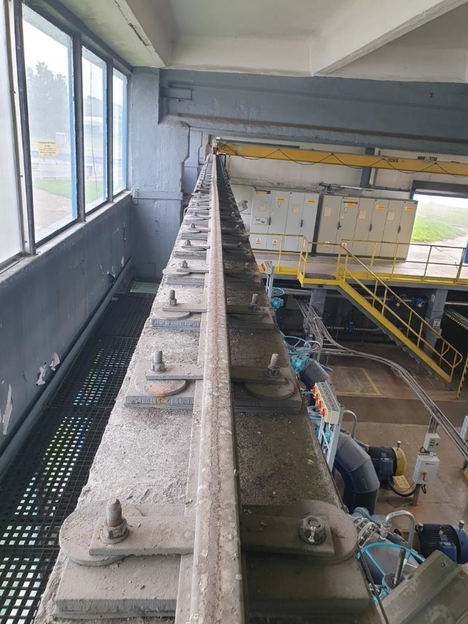 Wnętrze przemysłowe: widok z góry na betonową konstrukcję z metalowymi elementami mocującymi, rzędem okien po lewej stronie i żółtą metalową konstrukcją schodów w tle, ukazujący infrastrukturę...