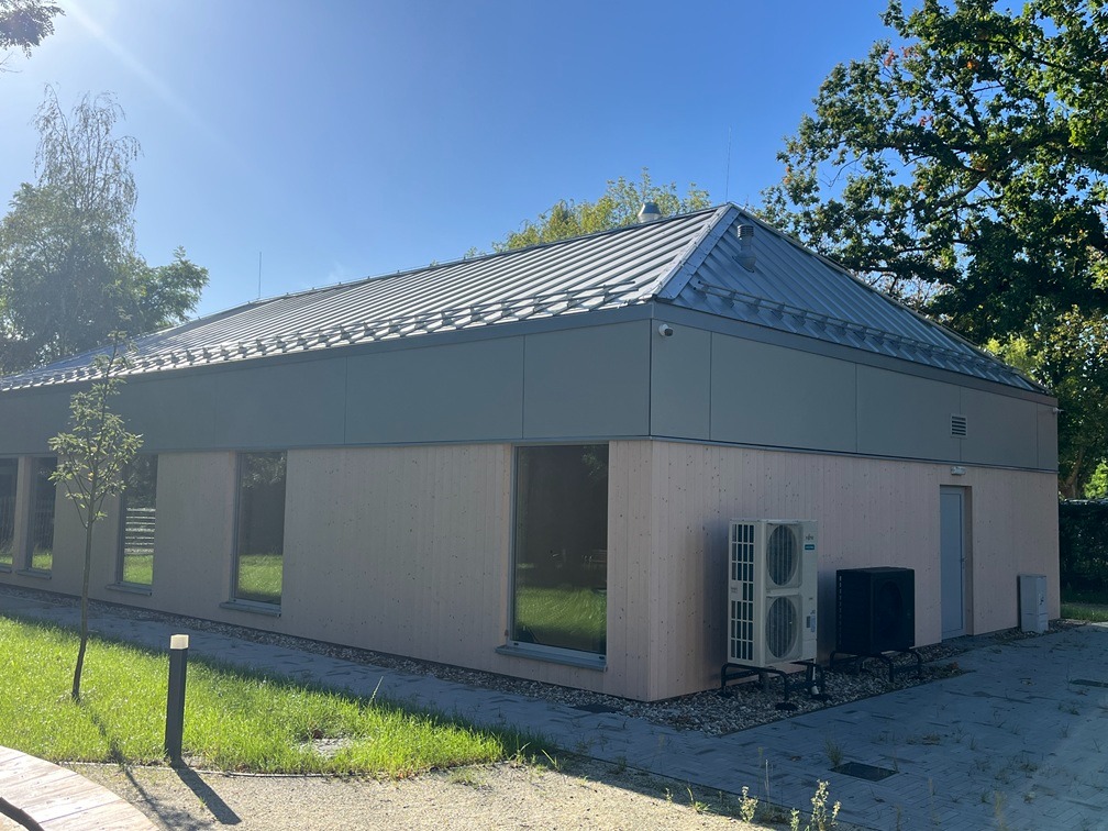 Nowoczesny budynek parterowy z jasną elewacją, szarym dachem i klimatyzacją, otoczony zielenią w słoneczny dzień. Widok z zewnątrz, prezentujący architekturę budynku.