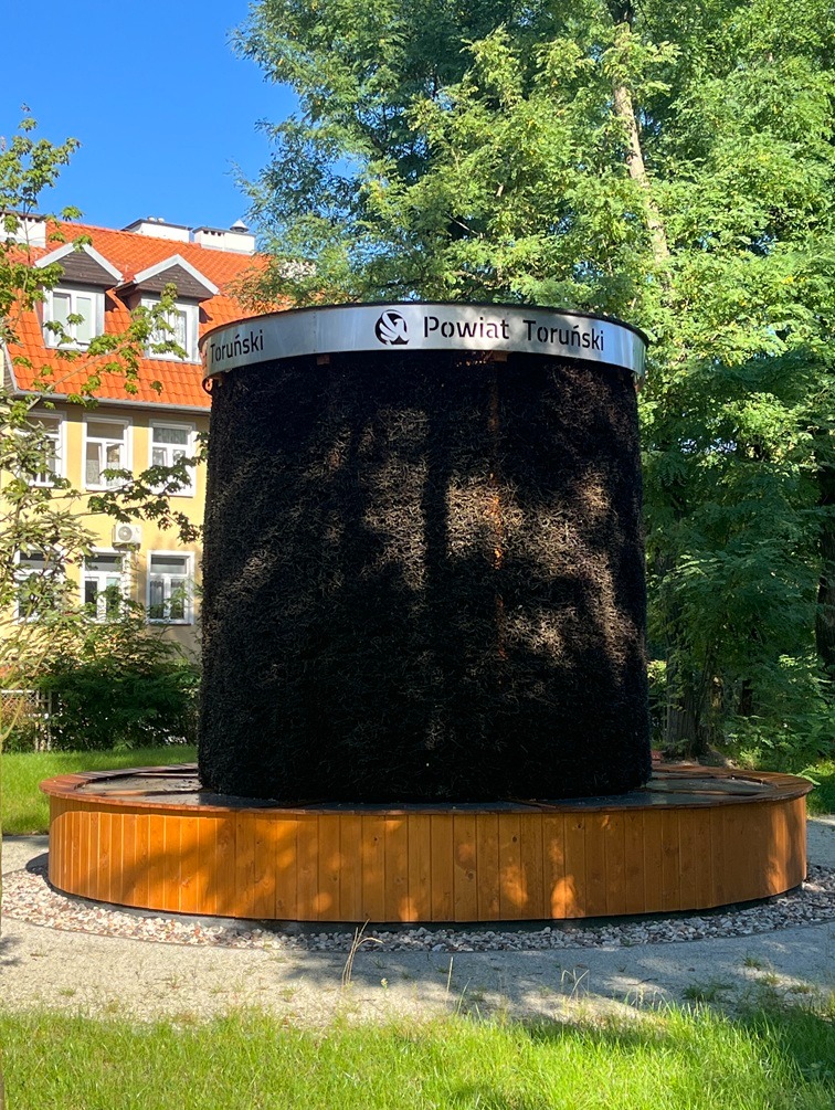 Tężnia solankowa w Toruniu, otoczona zielenią. Konstrukcja z drewna i tarniny z napisem 'Powiat Toruński'. W tle budynek z czerwonym dachem i drzewa.