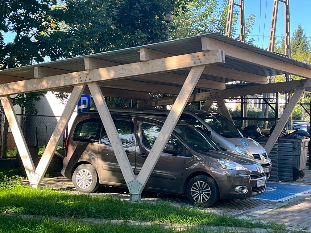 Drewniana wiata parkingowa z blachy falistej, wsparta na konstrukcji z belek. Dwa zaparkowane samochody, jeden brązowy, drugi srebrny. Znak niebieski dla niepełnosprawnych.
