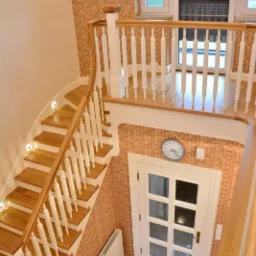 Klatka schodowa z drewnianymi schodami z podświetleniem LED, białymi balustradami i kryształowym żyrandolem, prowadząca do białych drzwi z przeszkleniami i widocznym zegarem na ścianie.