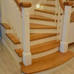 Drewniane schody z białymi balustradami i podświetleniem LED na ścianie, widoczne detale wykończenia i wzór na słupku.