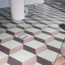 Wz&oacute;r 3D na podłodze w odcieniach szarości, r&oacute;żu i zieleni, z widocznymi otworami rewizyjnymi i kolumną w tle, częściowo zasłonięty przez szkło.