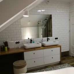 Jasna łazienka na poddaszu z dwoma umywalkami nablatowymi, drewnianym blatem, białymi szafkami i lustrem, w którym odbija się kabina prysznicowa z czarną armaturą.