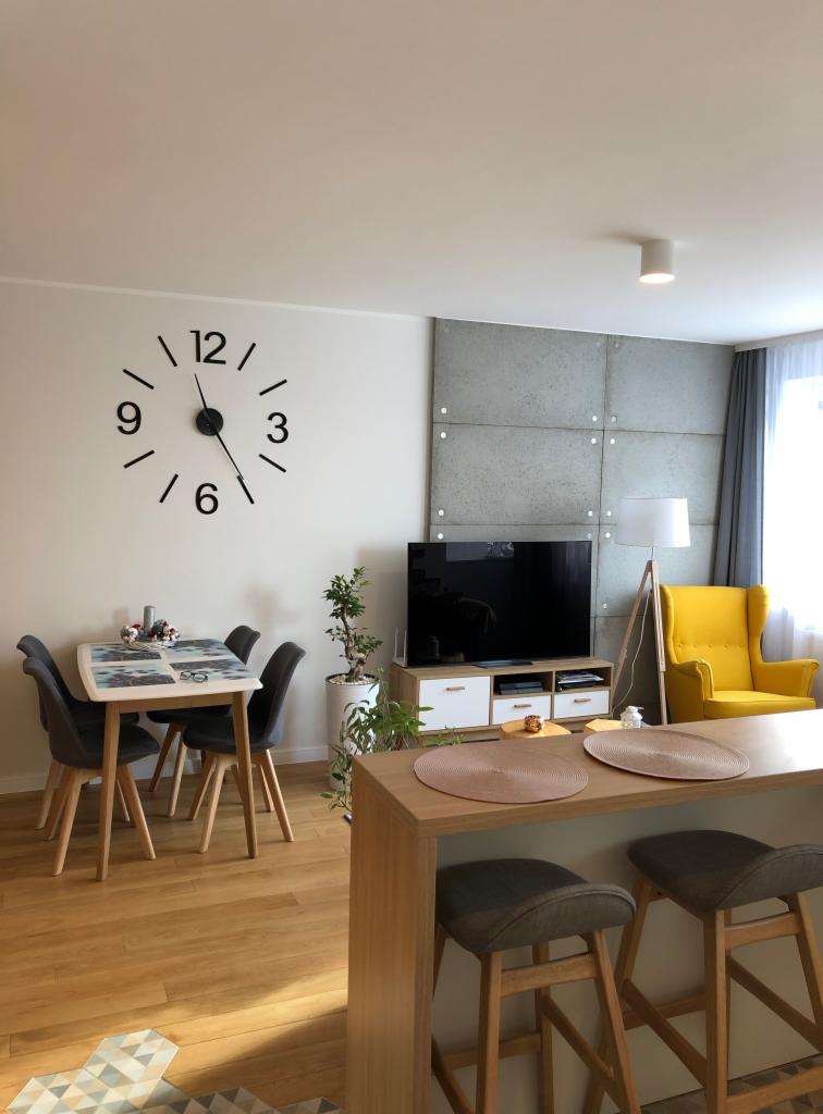 Jasne, nowocześnie urządzone wnętrze: stół z krzesłami, ściana z betonowych paneli, duży zegar ścienny, żółty fotel i drewniana podłoga z geometrycznym wzorem dywanu.