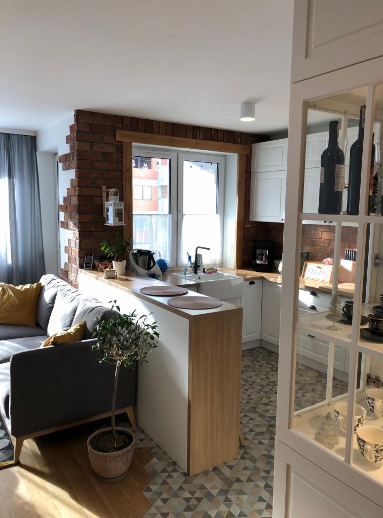 Salon z aneksem kuchennym: ceglana ściana, białe szafki, drewniany blat, szara sofa z żółtymi poduszkami i dekoracyjna witryna z butelkami wina.
