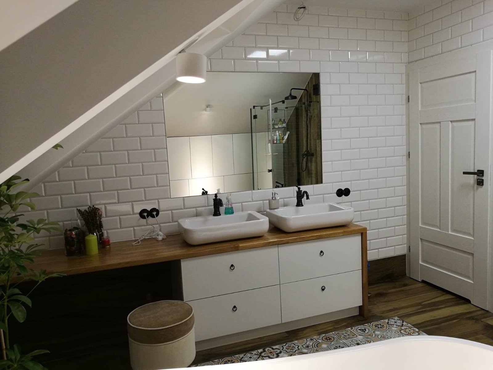 Jasna łazienka na poddaszu z dwoma umywalkami nablatowymi, drewnianym blatem, białymi szafkami i lustrem, w którym odbija się kabina prysznicowa z czarną armaturą.