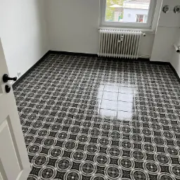 Ukończona podłoga z wzorzystych, czarno-białych płytek ceramicznych w pustym pomieszczeniu z grzejnikiem pod oknem i częściowo otwartymi białymi drzwiami.
