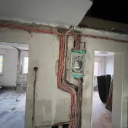 Instalacja elektryczna w trakcie remontu: kable prowadzone po ścianie, puszka z przewodami zabezpieczona pianką, surowy stan ścian i widoczne przejścia do innych pomieszczeń.