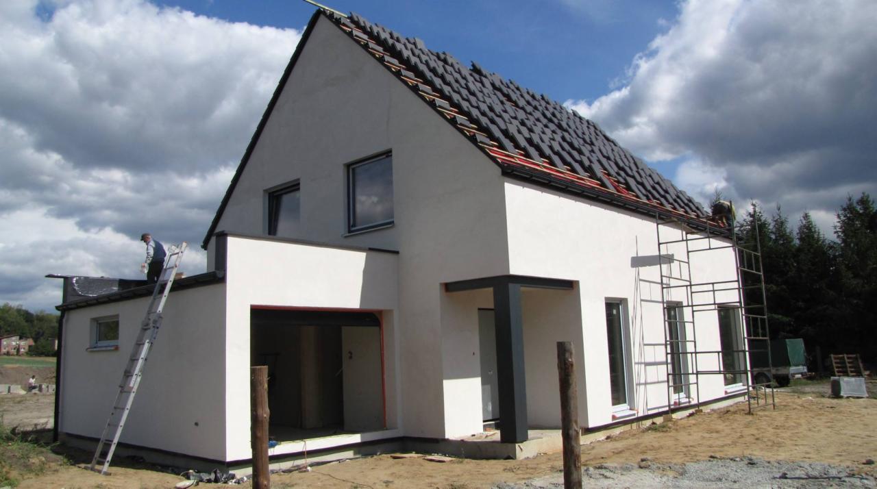 Montaż dachówki na dachu nowoczesnego domu w trakcie budowy, widoczne rusztowania i pracownicy.