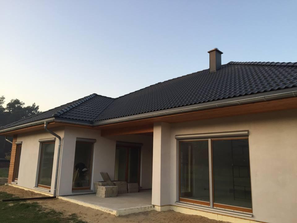 Nowoczesny parterowy dom z szarą dachówką, drewnianą podbitką i zewnętrznymi roletami, widok z boku na taras.