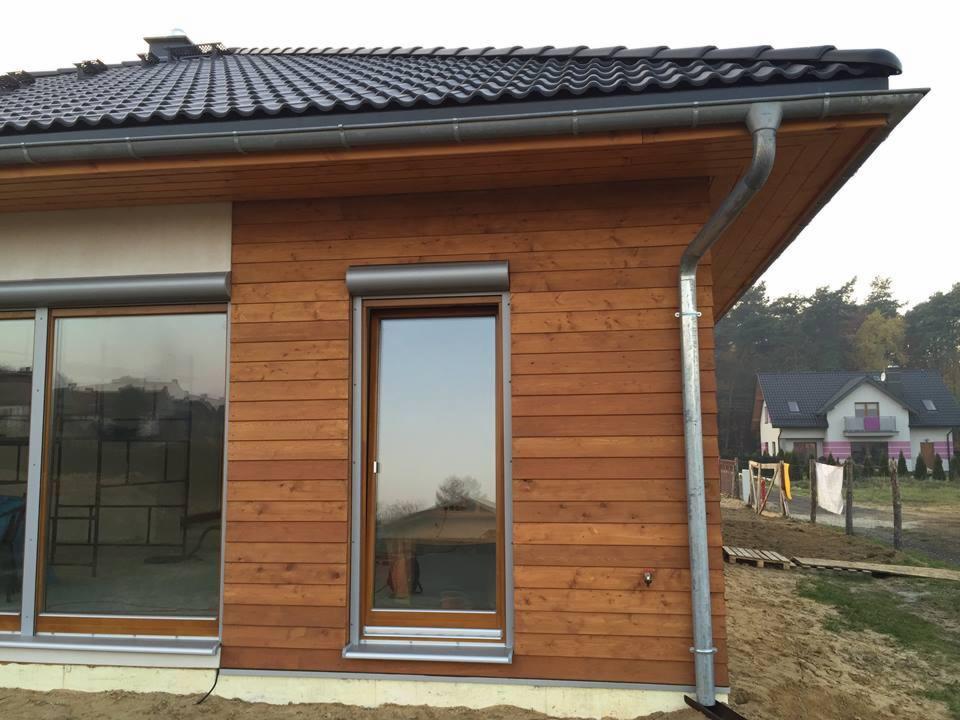 Fragment domu szkieletowego z drewnianą elewacją, roletami zewnętrznymi i systemem rynnowym, widok z zewnątrz na tle innego domu i drzew