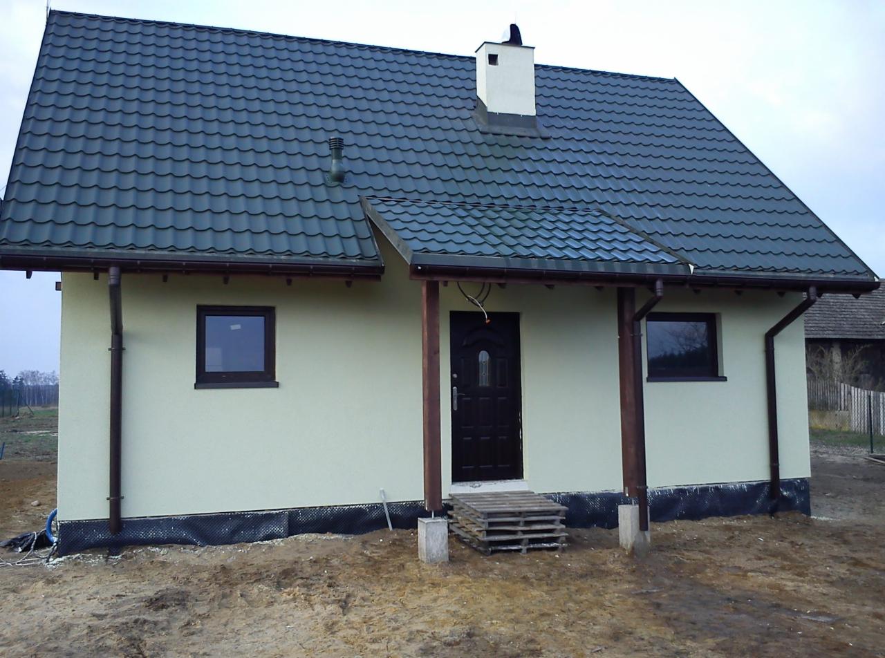 Gotowy dom szkieletowy z ciemnozielonym dachem z blachodachówki, jasną elewacją i drewnianą wiatą nad wejściem, widoczne palety przed drzwiami wejściowymi.