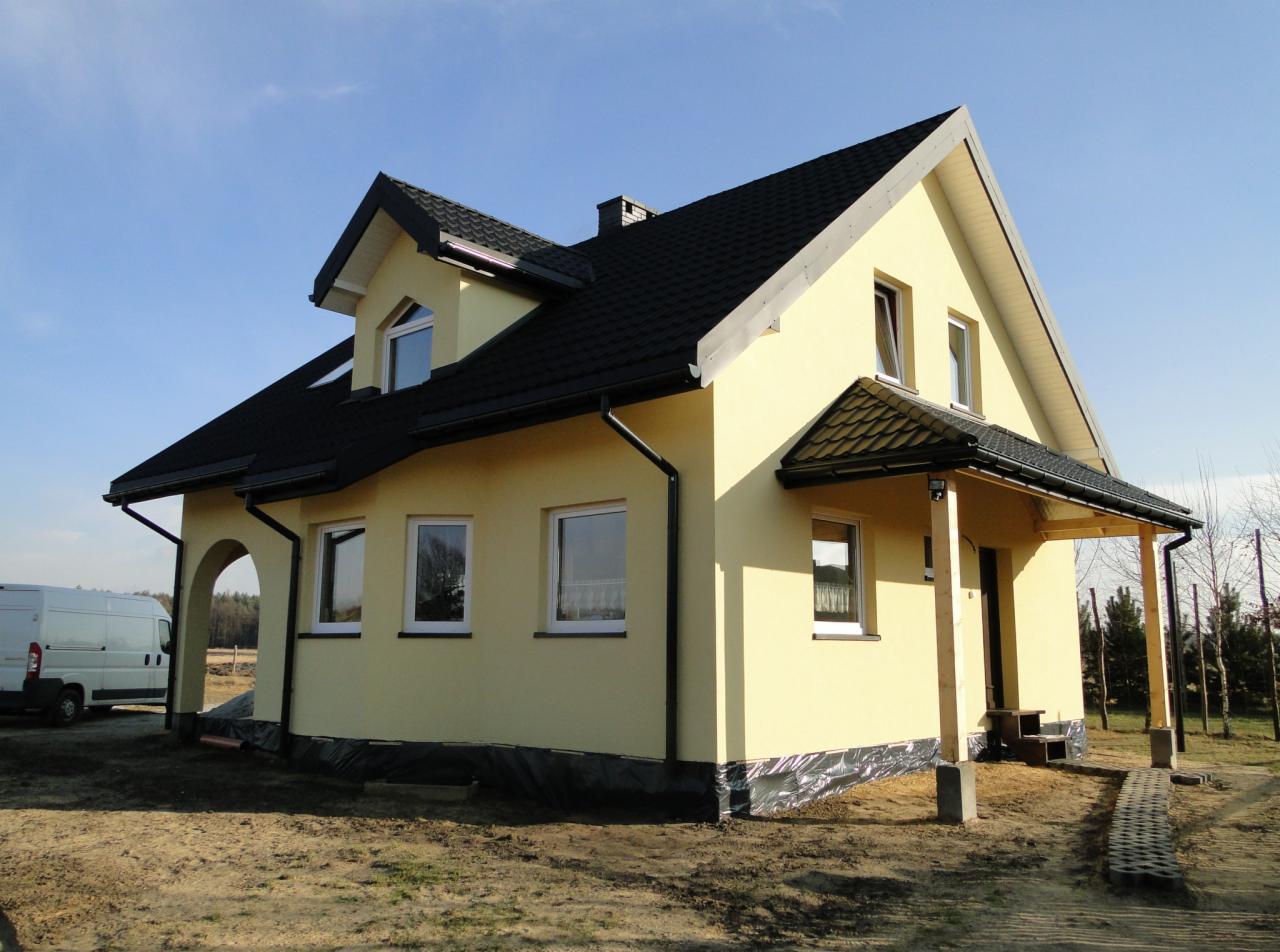 Żółty dom z czarnym dachem w trakcie wykończenia, widoczna folia ochronna na fundamencie, podcień wejściowy z surowego drewna, biały van w tle.