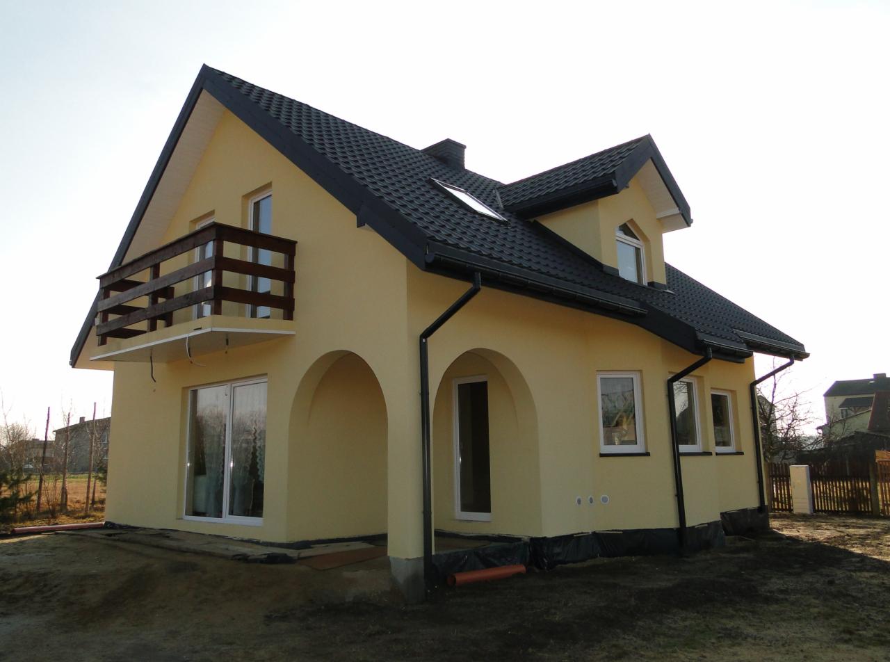 Żółty dom jednorodzinny z czarnym dachem, balkonem z drewnianą balustradą i łukowym wejściem, otoczony przygotowanym terenem pod ogród.