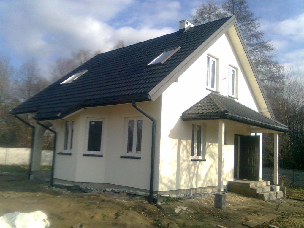 Gotowy, parterowy budynek z czarną dachówką, z oknami dachowymi i małym gankiem, w surowym otoczeniu.