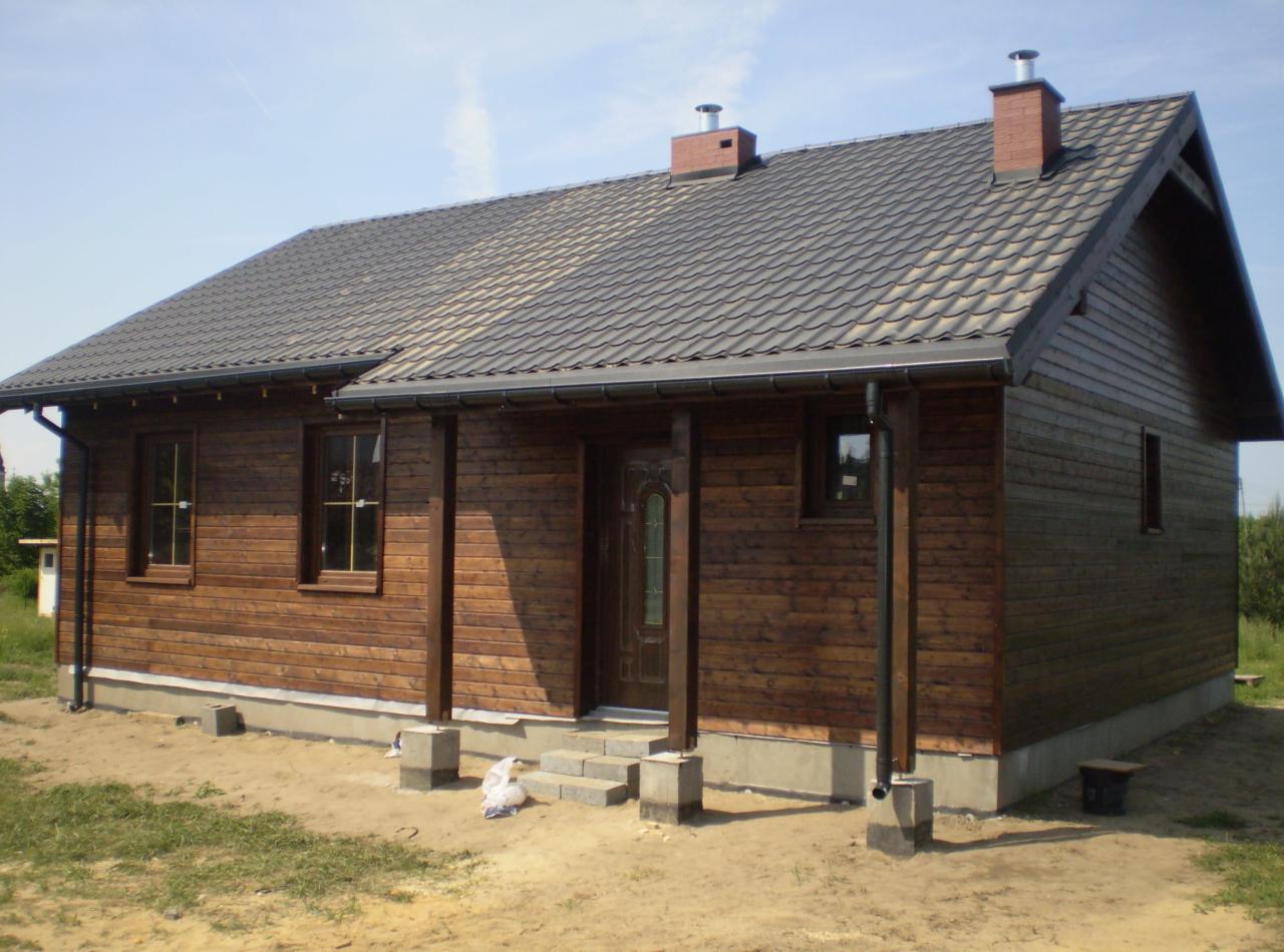 Niski, parterowy budynek z drewnianą elewacją w odcieniach brązu, z ciemnoszarą dachówką, widoczny z zewnątrz w słoneczny dzień.