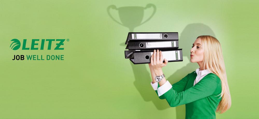 Kobieta z blond włosami w zielonej bluzce trzyma trzy czarne segregatory biurowe marki Leitz, na zielonym tle z cieniem pucharu.