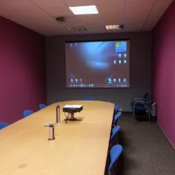 Sala konferencyjna z projektorem wyświetlającym ekran komputera, na długim drewnianym stole widoczne gniazda zasilające i urządzenie do wideokonferencji.