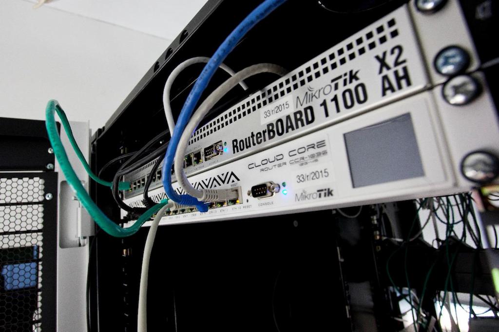 Szczegółowe ujęcie szafy rack z zainstalowanymi routerami MikroTik RouterBOARD 1100 AH i Cloud Core Router CCR-1036, widoczne kable sieciowe w kolorach niebieskim, białym i zielonym podłączone...