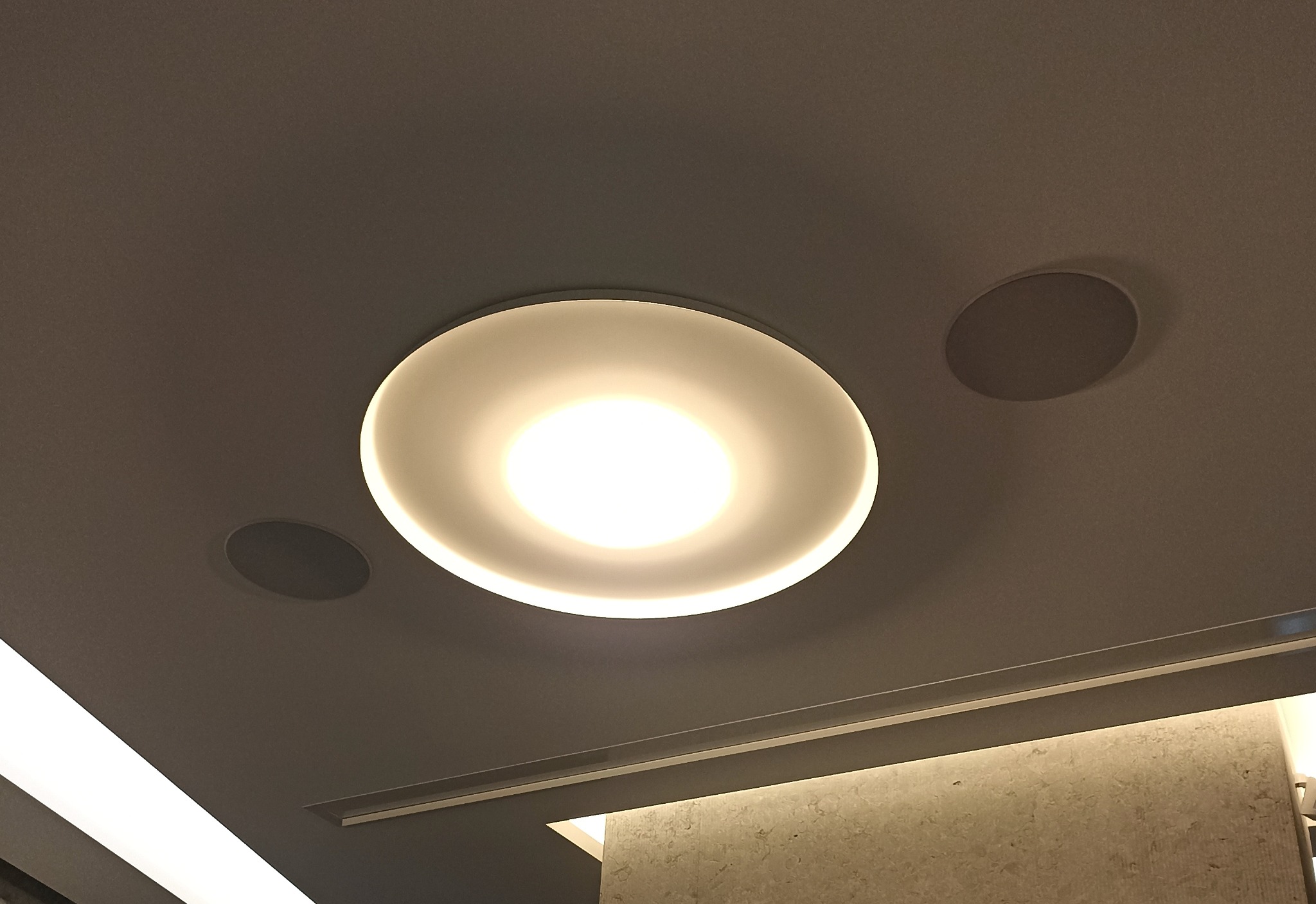 Okrągła, podwieszana lampa sufitowa LED włączona, z dwoma okrągłymi głośnikami w tle, oświetlenie dekoracyjne wzdłuż krawędzi sufitu.