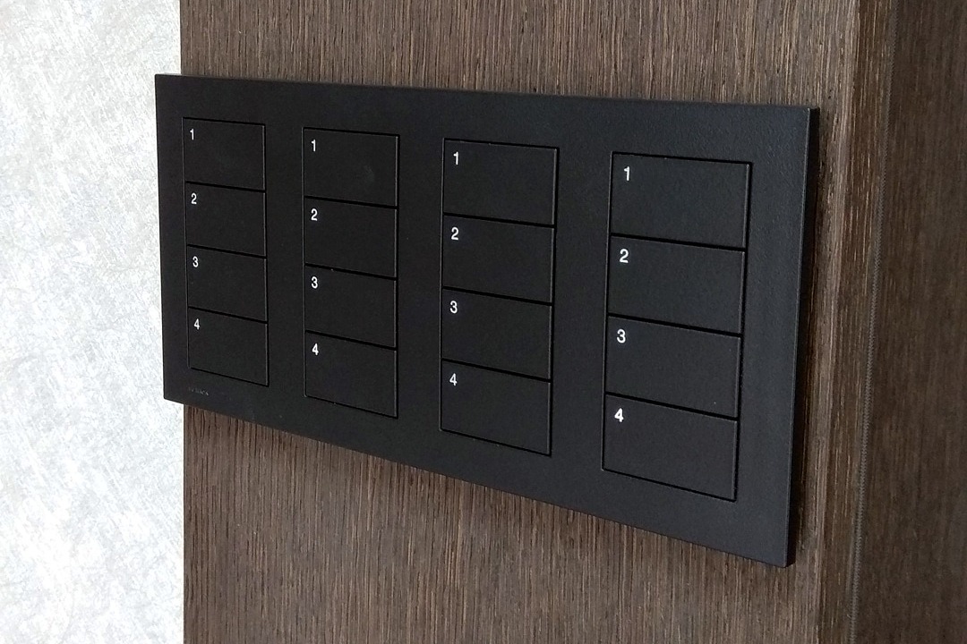 Czarny panel sterowania z czterema rzędami przycisków oznaczonych cyframi 1-4, zamontowany na drewnianej ścianie w nowoczesnym wnętrzu.