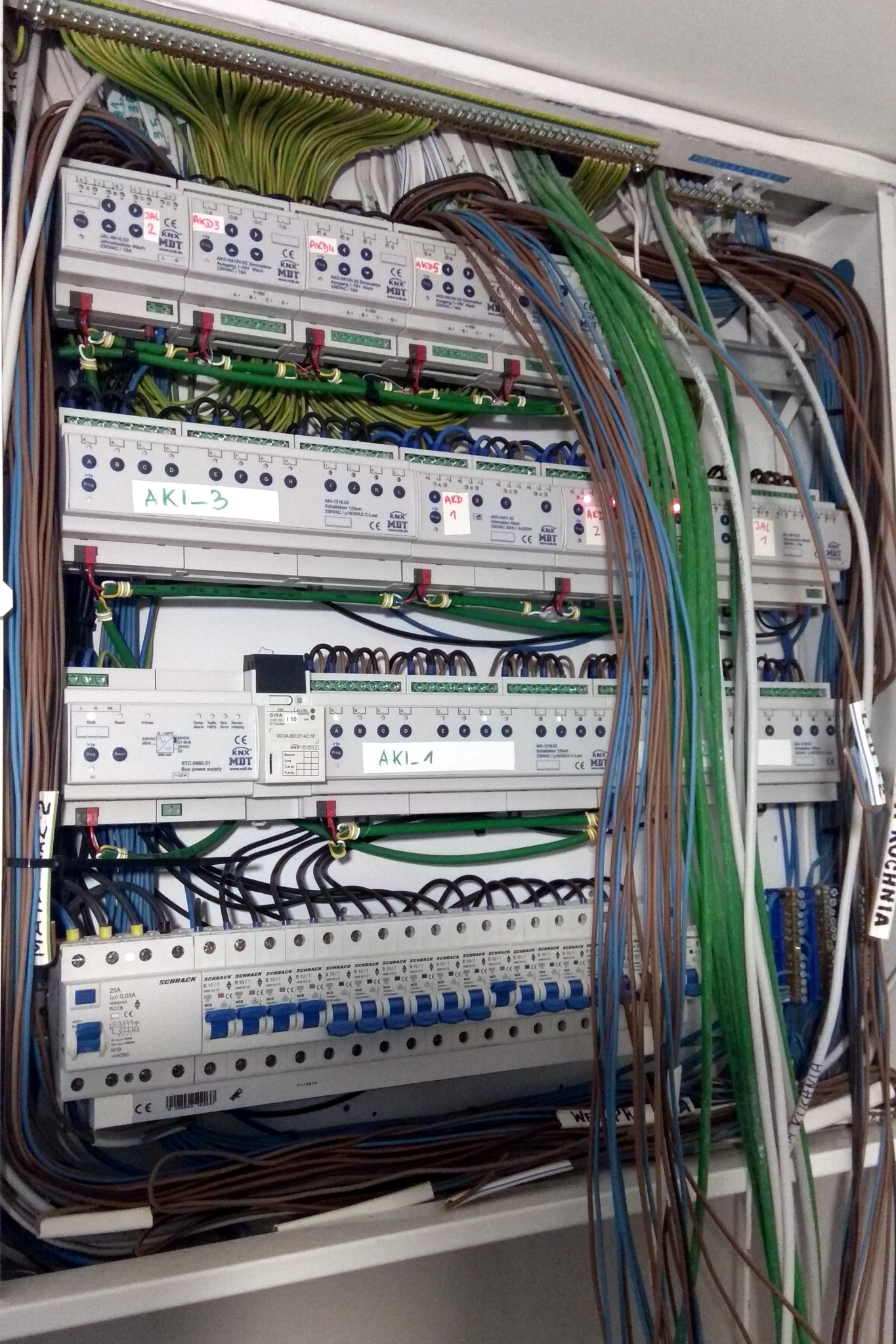 Otwarta szafa rozdzielcza z modułami sterującymi inteligentnego domu, widoczne liczne przewody i moduły KNX MDT oraz Schrack z opisami AKI-1, AKI-3, AKD1 i AKD3.