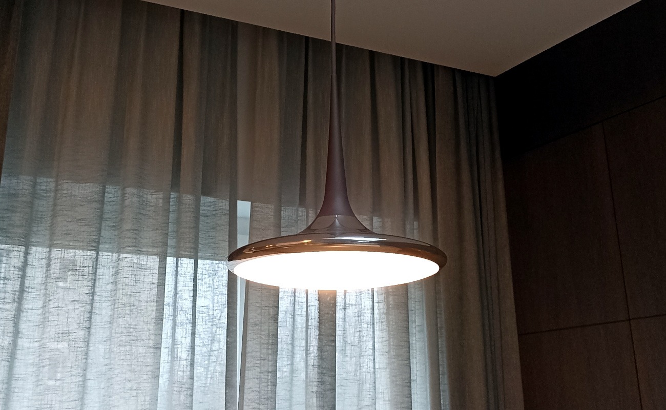 Nowoczesna lampa wisząca z okrągłym, podświetlanym kloszem, zawieszona na tle zasłoniętego okna i ciemnej, drewnianej ściany.