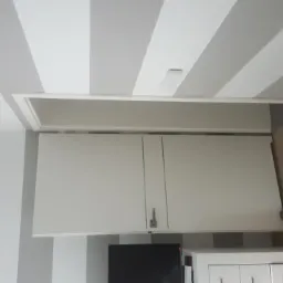 Wnętrze po odświeżeniu: ściany i sufit w szaro-białe pasy, widoczny fragment zabudowy kuchennej, na podłodze puszka z farbą i taśma malarska.