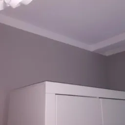 Ściana w odcieniach szarości z białą listwą przysufitową, widoczny fragment białej szafy z przesuwnymi drzwiami i lampa wisząca.