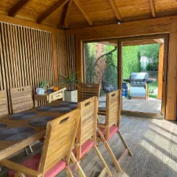 Drewniana altana z drewnianymi meblami ogrodowymi, widok na grill i wiszące krzesło przez otwarte drzwi przesuwne.