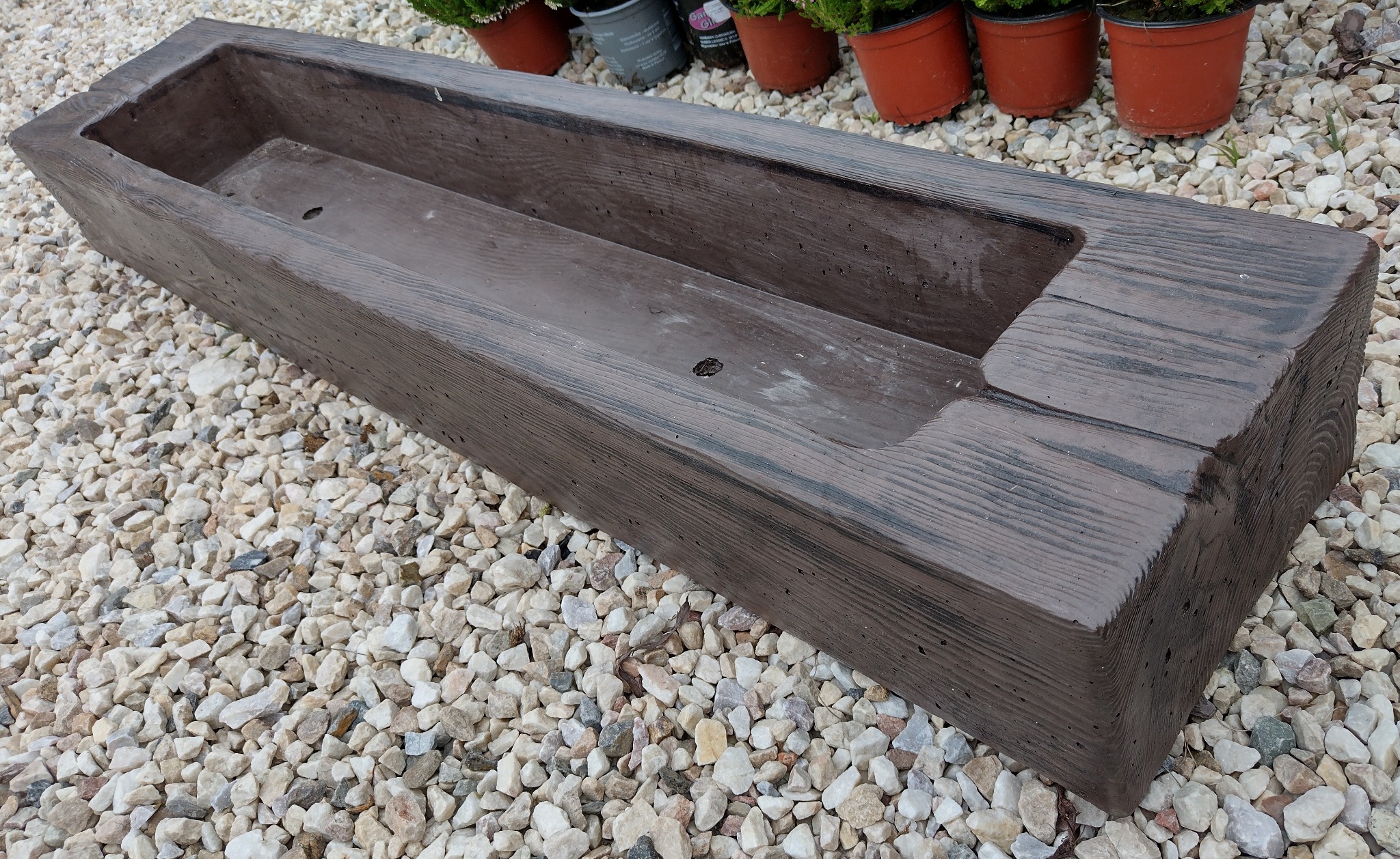 Podłużna, brązowa donica z betonu imitującego drewno, ustawiona na żwirowym podłożu, z widocznymi śladami po szalunku i nierówną fakturą powierzchni.