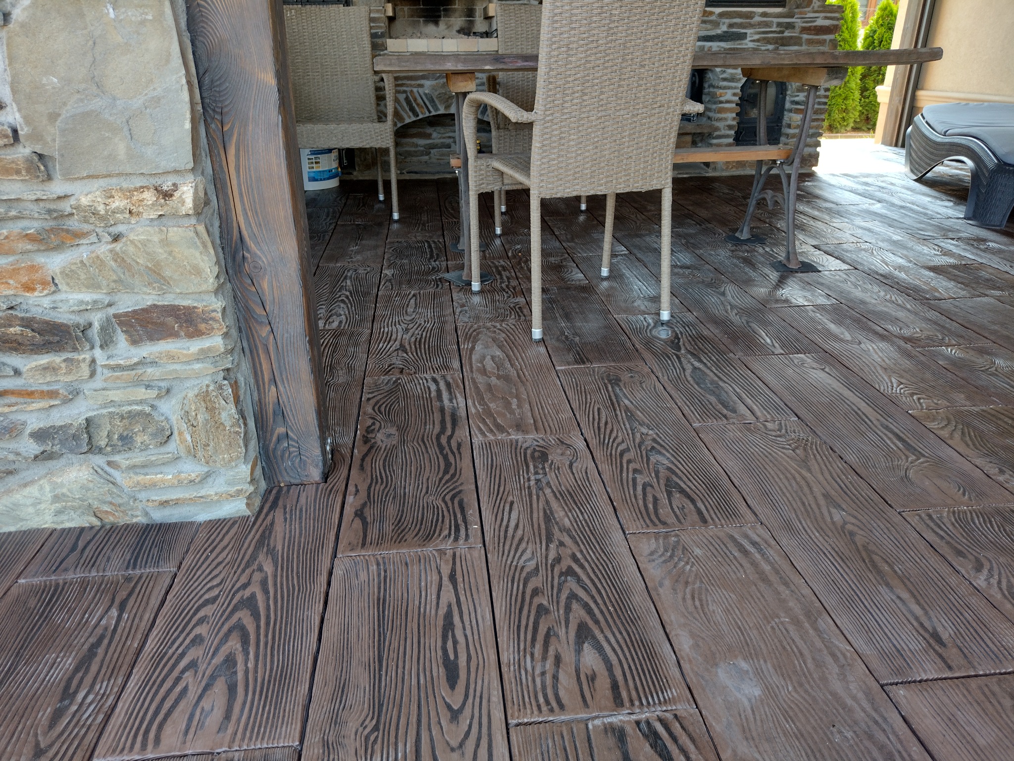 Zewnętrzna powierzchnia tarasu wyłożona brązową nawierzchnią imitującą deski, z widocznym kamiennym filarem i meblami ogrodowymi.