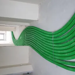 Zielone rury wentylacyjne karbowane przymocowane do sufitu i ściany w surowym stanie, widoczne okno i kable elektryczne.